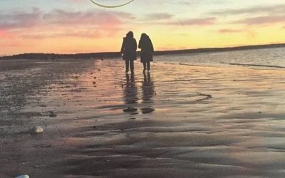 Two people walk on a wet beach at sunset,