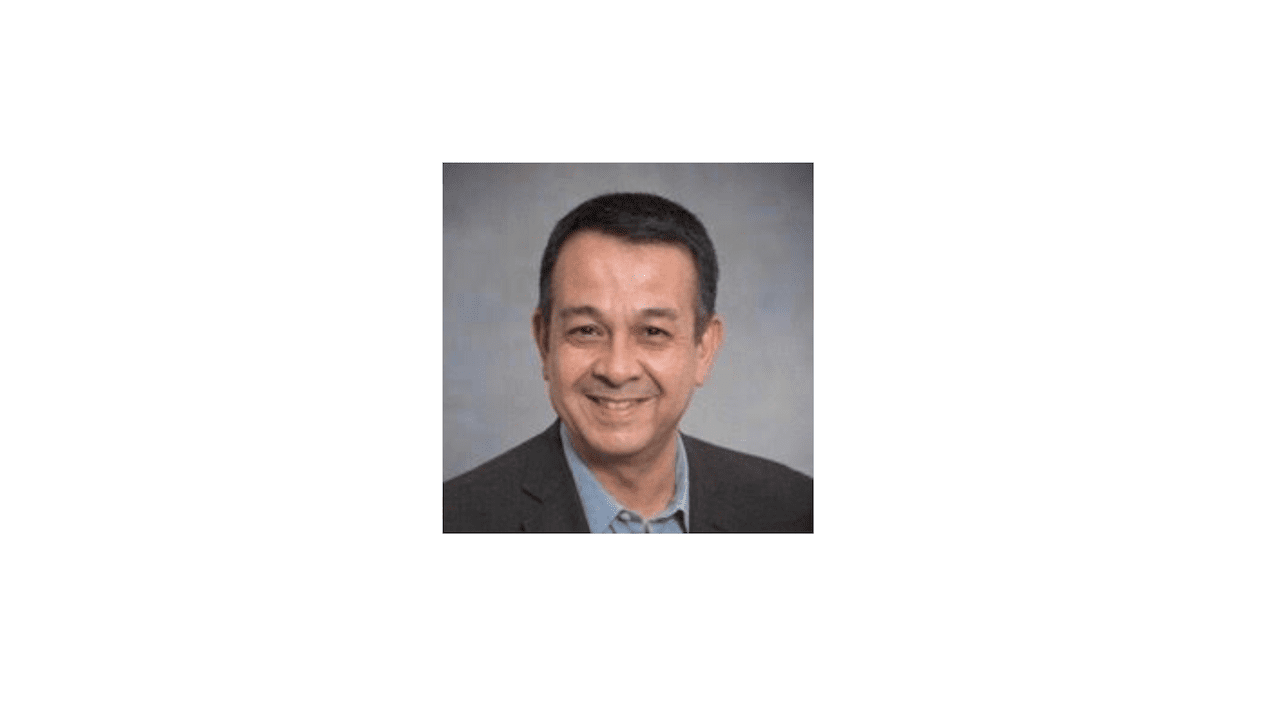 Smiling man with dark hair and a blue shirt against a grey background.