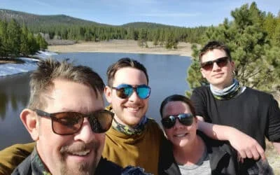 Four smiling people wearing sunglasses outdoors by a lake with