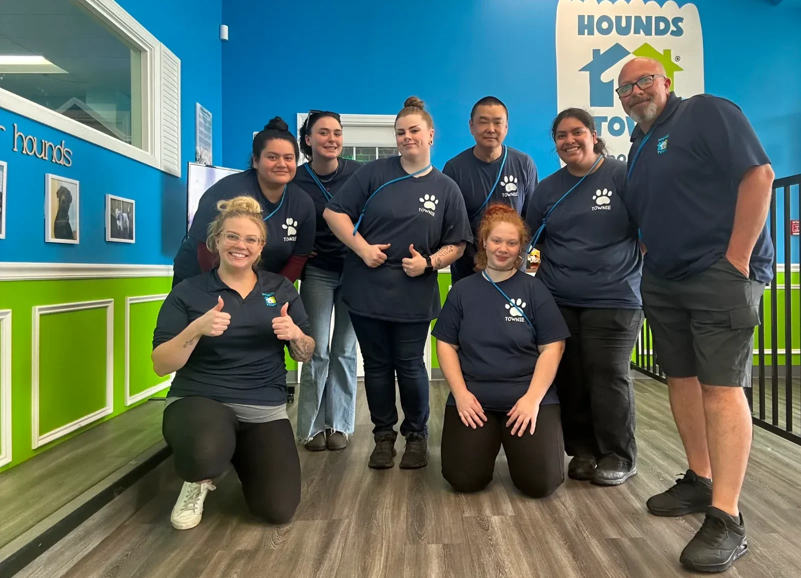 A group of smiling staff members at Hounds Town dog daycare, wearing matching shirts and giving thumbs up.