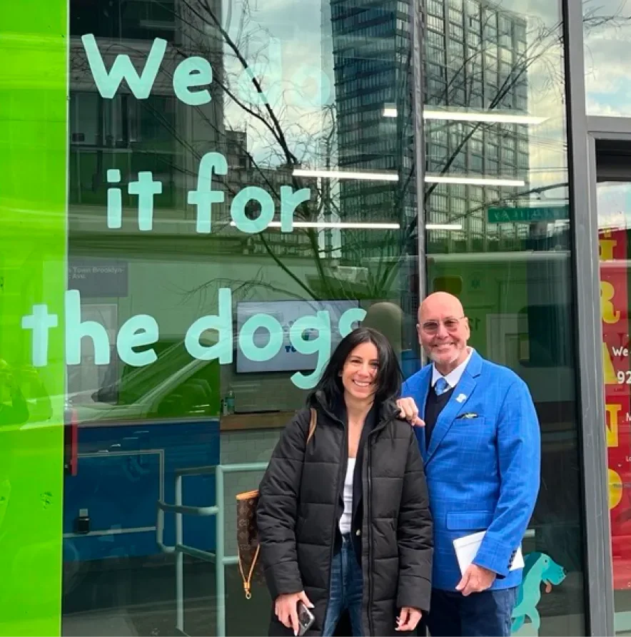Two people stand in front of a colorful storefront with the phrase "We do it for the dogs" displayed prominently.