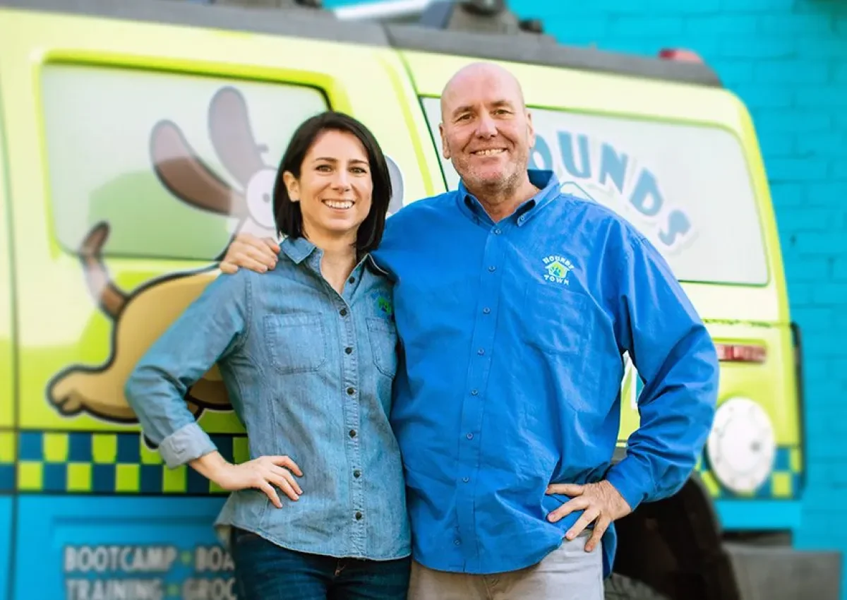 Two smiling people stand in front of a colorful dog daycare van, promoting a dog daycare franchise opportunity.