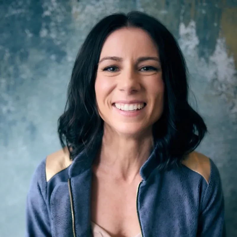 A smiling woman with dark hair wearing a blue jacket against a textured background.